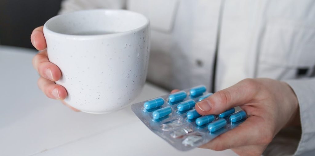 Человек держит капсулы в одной руке, а в другой - кружку с лекарством.








