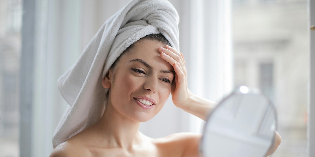 Woman with a towel on her head looking at her face in the mirror.