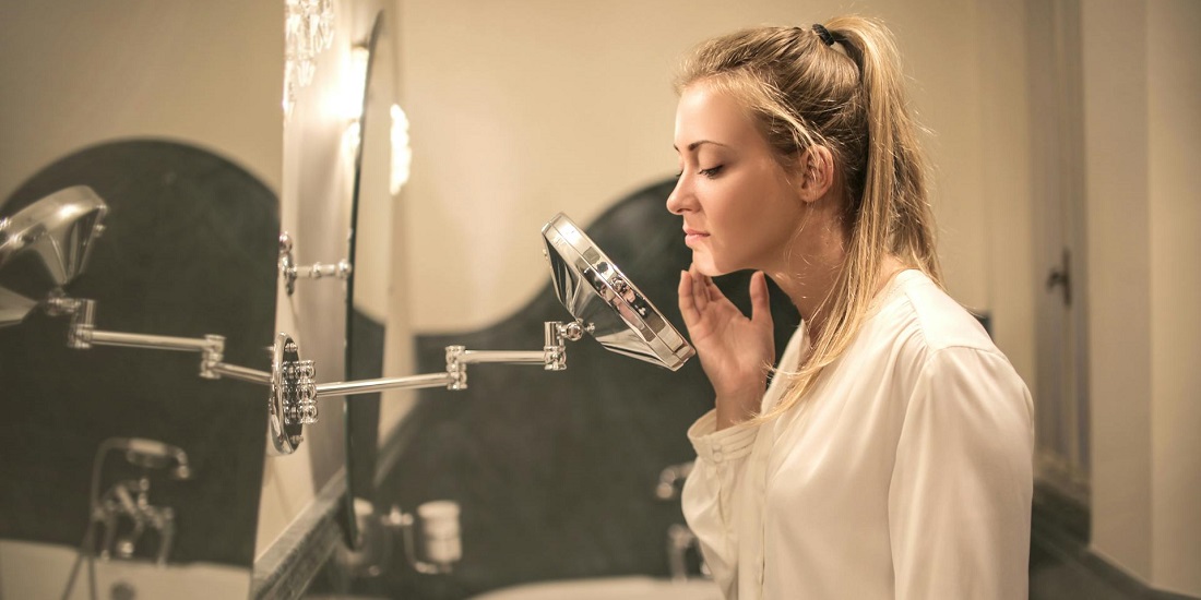 Mladu ženu u beloj košulji zanima stanje kože dok stoji ispred ogledala