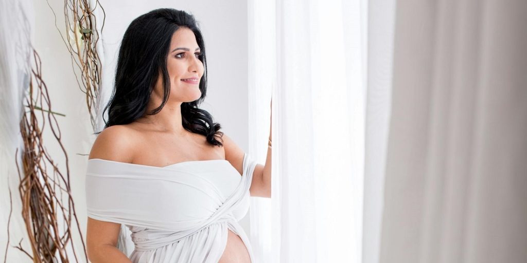 A smiling pregnant woman in a white dress is looking through the window