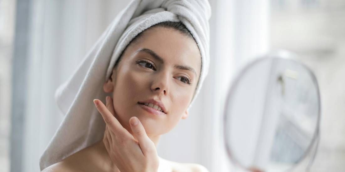 A woman with a towel on her head looks at herself in the mirror and examines the results achieved with azelaic acid.