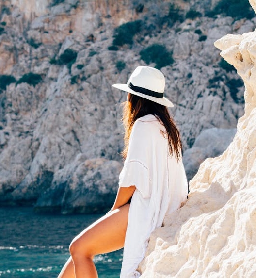 Osoba u letnjoj odeći i šeširu sedi na steni pored mora